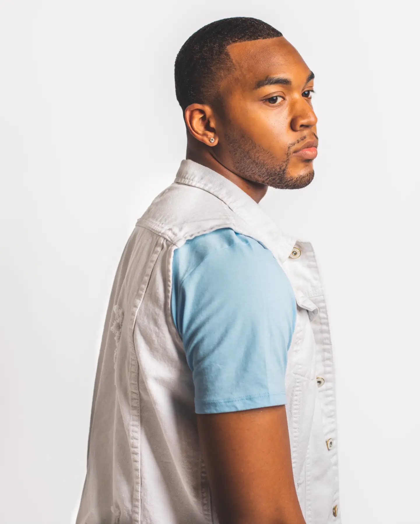 Studio portrait of Bren Got Hits in a white denim vest over a light blue tee, looking over his shoulder